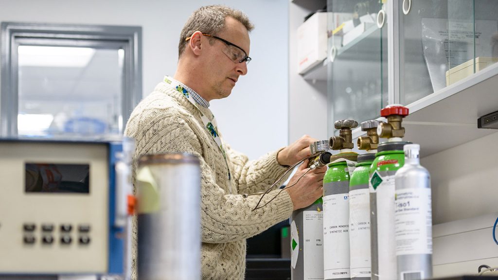 Man wearing beige jumper and safety glasses applies labels to gas canisters on a work bench