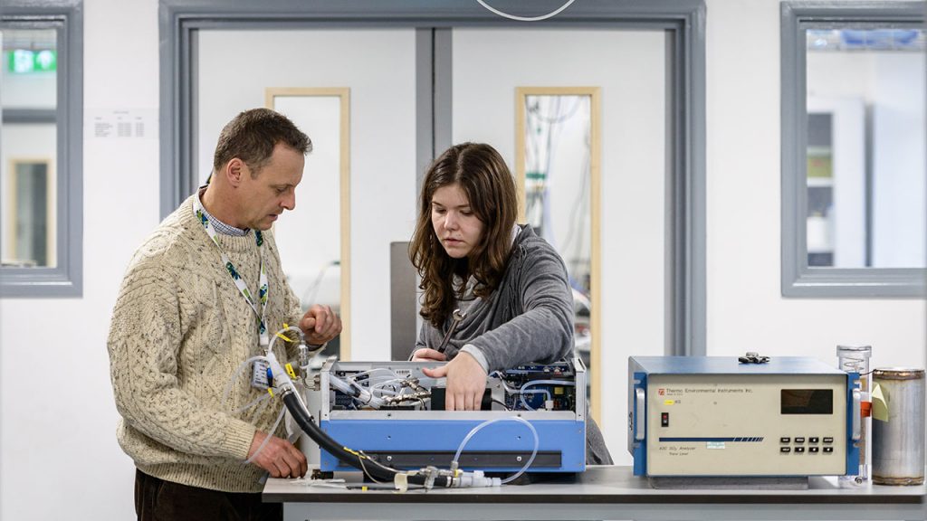 Man wearing beige knitted jumper and female wearing grey jumper stand at a desk, inspecting a blue machine with wires.
