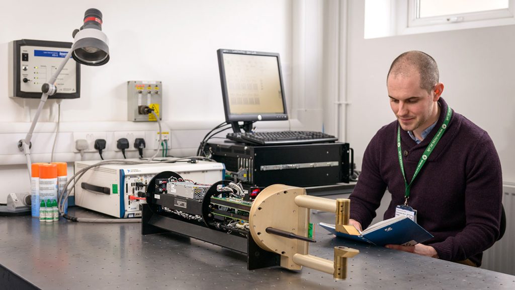 Man with shaved head wearing purple jumper reads blue book while sat at desk in laboratory