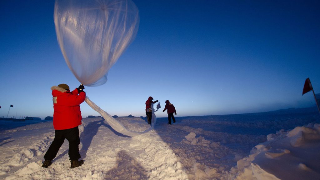 Person wearing thick orange coat holds a large balloon filled with helium, they stand on snowy ground.