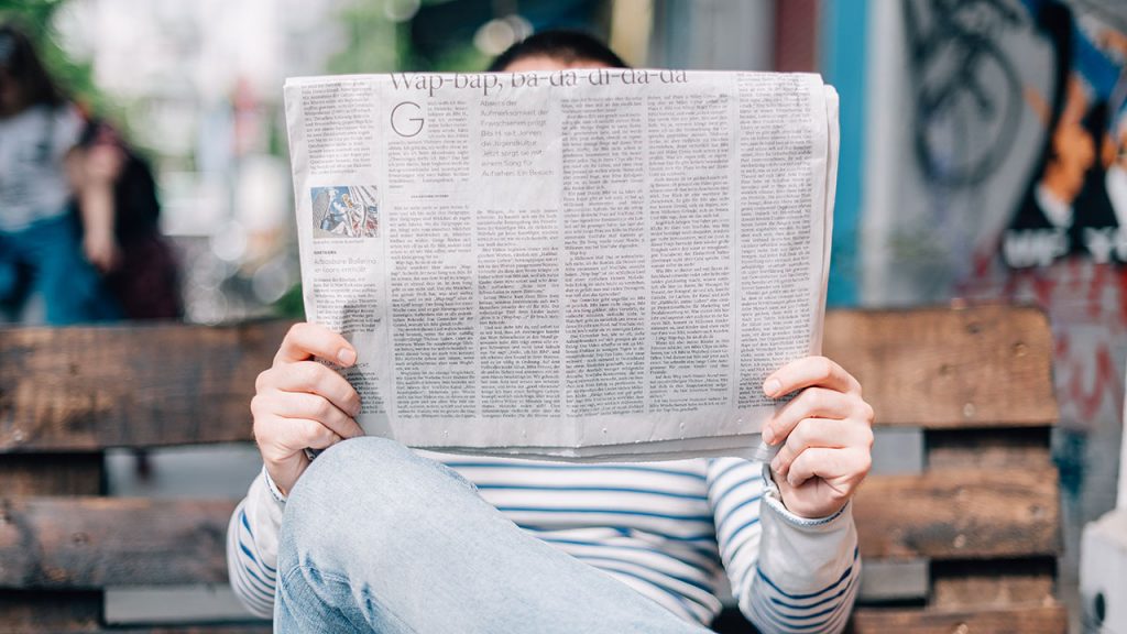 Man wearing jeans sits cross-legged and reads a large broadsheet newspaper