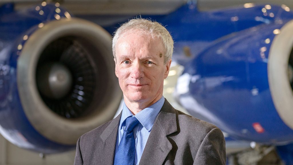 Professor Stephen Mobbs stands, wearing a grey suit, in front of blue aircraft engines